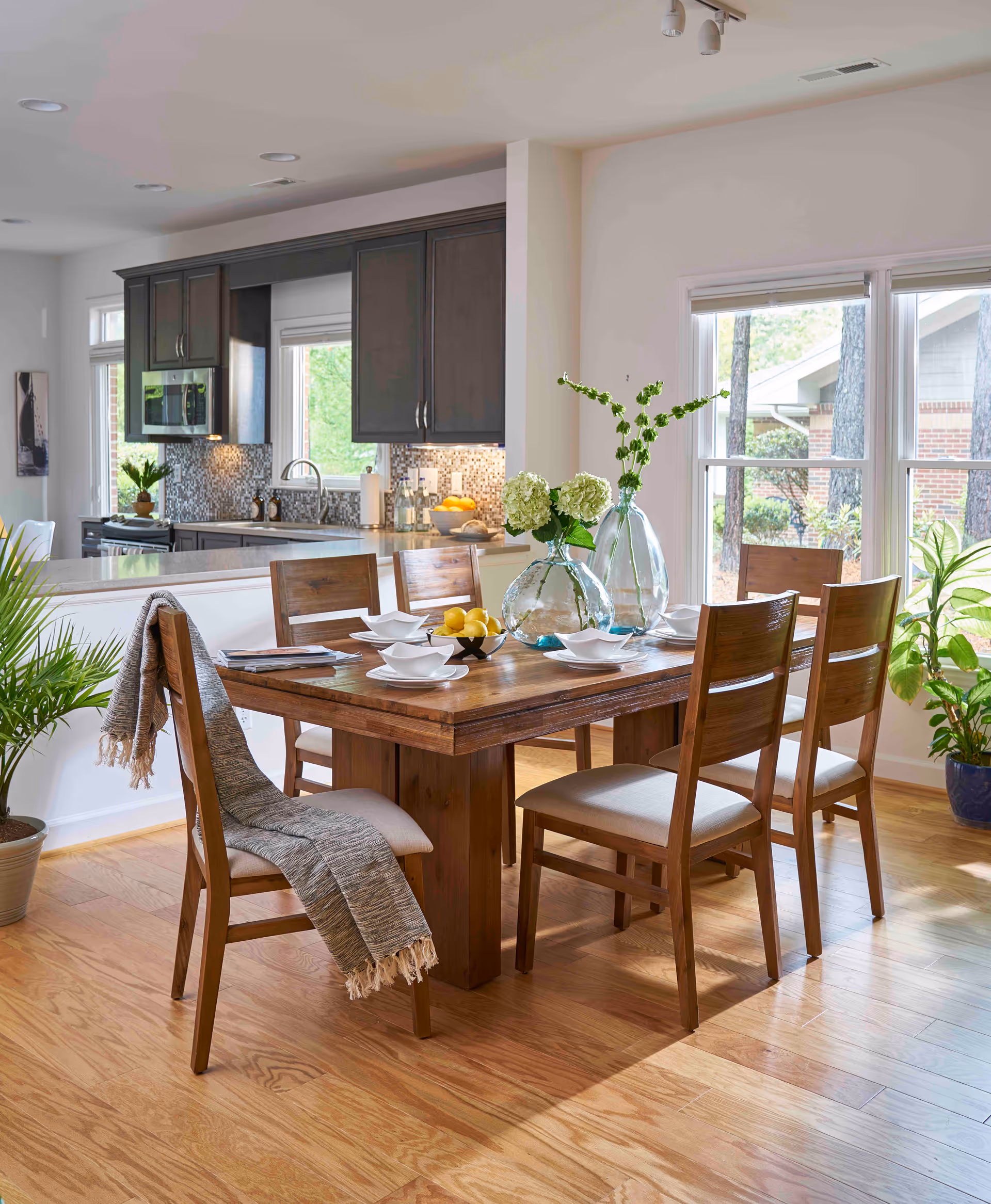Sunlit open-plan dining area with a wooden table set for four, decorative vases and a view into a modern kitchen.