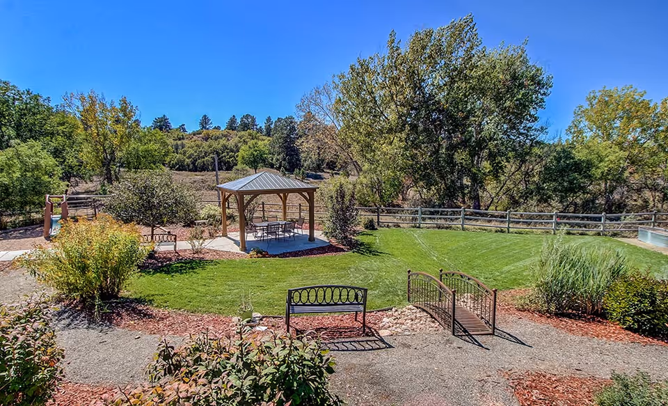 A well-maintained outdoor garden area with a small wooden gazebo containing a table and chairs, surrounded by green grass, trees, shrubs, and a wooden fence in the background under a clear blue sky. There is a small decorative bridge and a bench in the foreground.