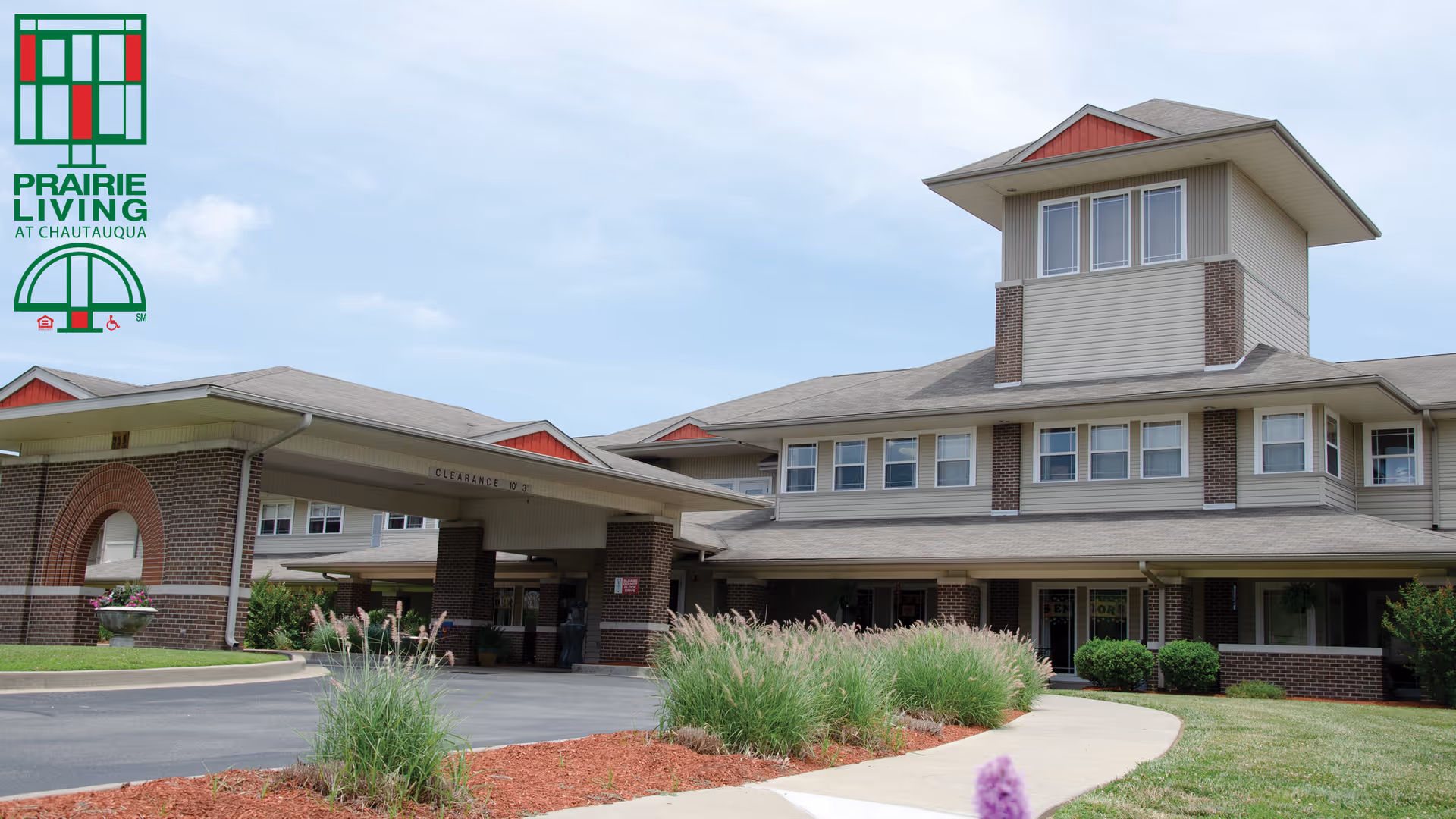 Exterior view of Prairie Living at Chautauqua facility showing a multi-story building with a covered entrance, brick and siding facade, landscaped greenery, and a clear sky.