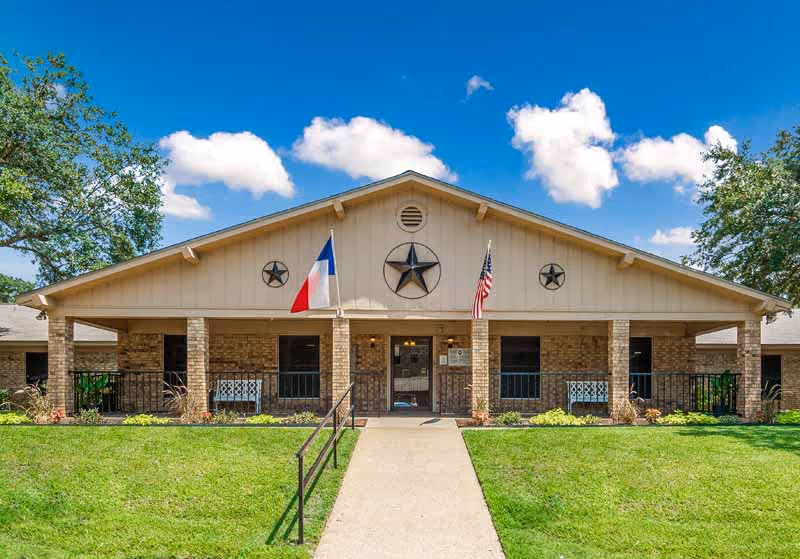 Front exterior view of Park Highlands Nursing & Rehabilitation Center building with a beige facade, brick pillars, two flags (Texas state flag and American flag), three star decorations on the upper wall, a concrete walkway leading to the entrance, and green grass lawn under a blue sky with some clouds.
