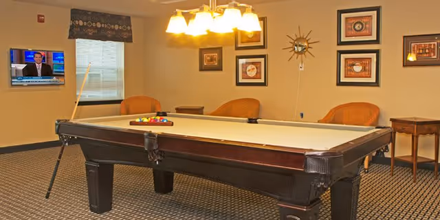 A recreation room with a pool table in the center, a wall-mounted TV, chairs, and framed artwork on the walls.