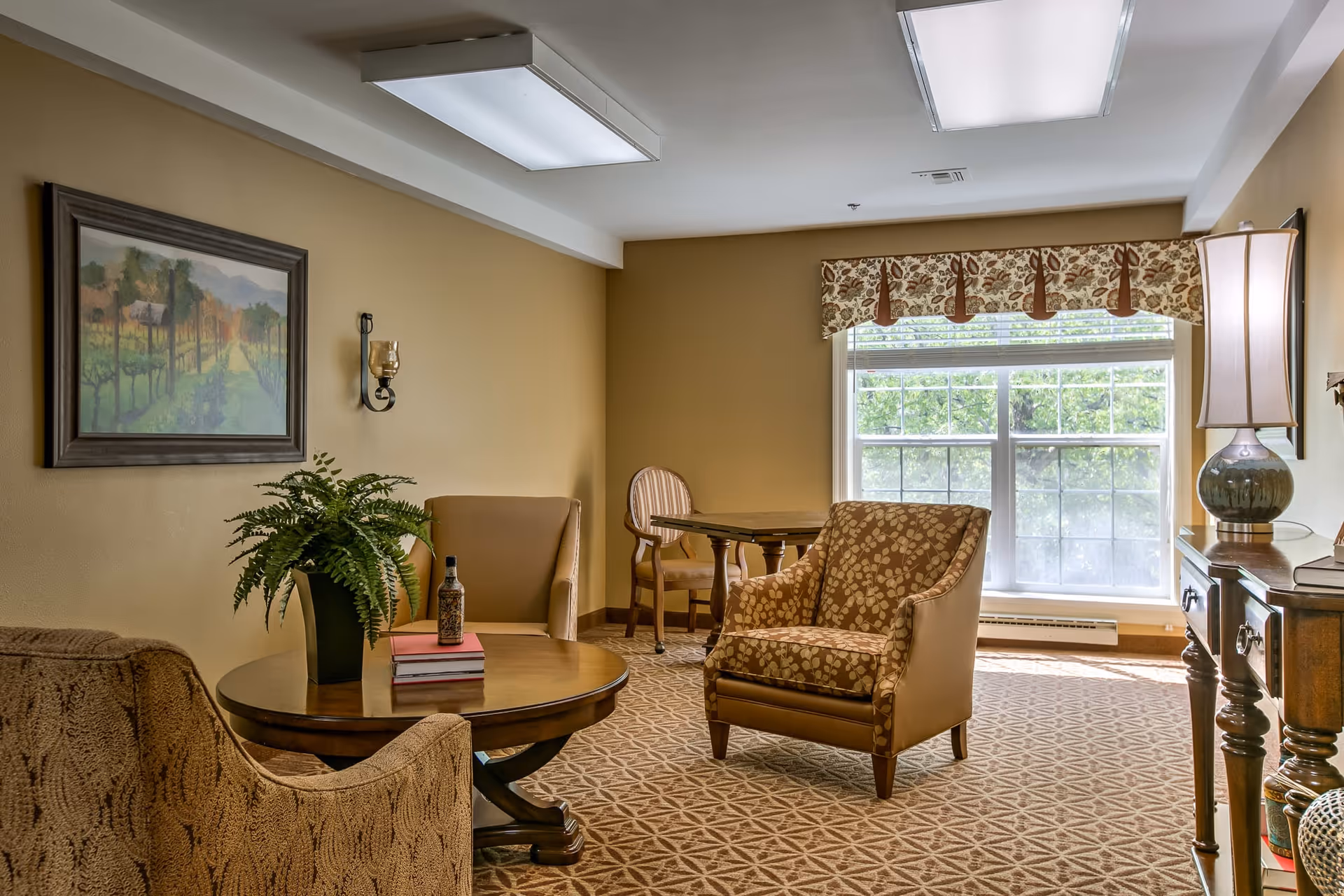 A cozy sitting area in a retirement community with patterned armchairs, a round wooden coffee table with a potted plant and books, a small wooden table with two chairs near a large window with floral valance, a framed landscape painting on the wall, and a wooden side table with a lamp and books.