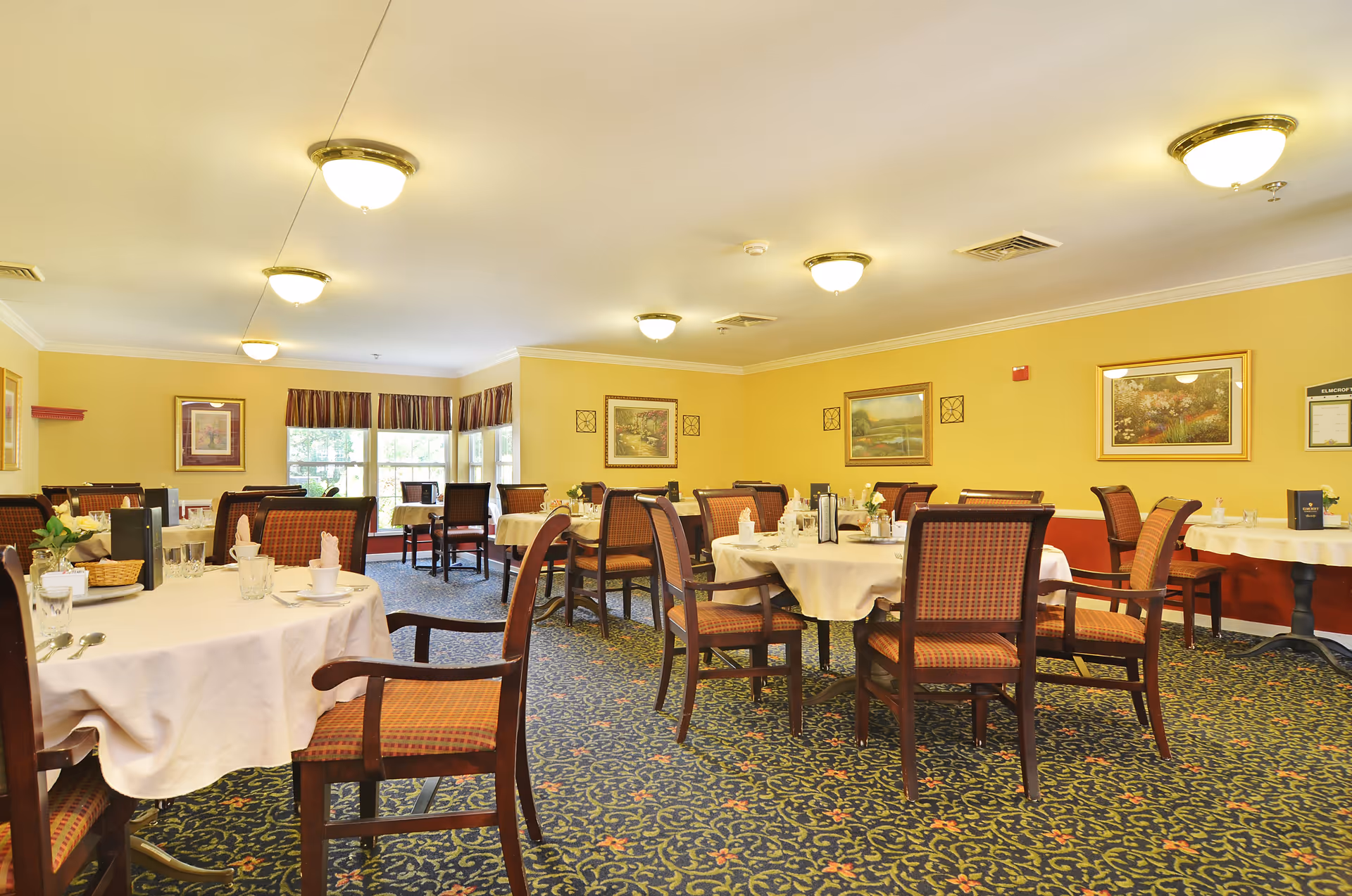 A dining room with round tables covered in white tablecloths, set with glasses, cups, and napkins. The room has yellow walls adorned with framed paintings and a patterned carpet. Several wooden chairs with cushioned seats are arranged around the tables. Ceiling lights provide warm illumination, and windows with valances allow natural light to enter.