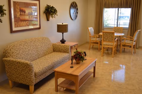 A cozy living area in a senior living facility featuring a patterned sofa, a wooden coffee table with a floral centerpiece, a side table with a lamp, a wall clock, framed artwork, and a round dining table with four chairs near a window with curtains.
