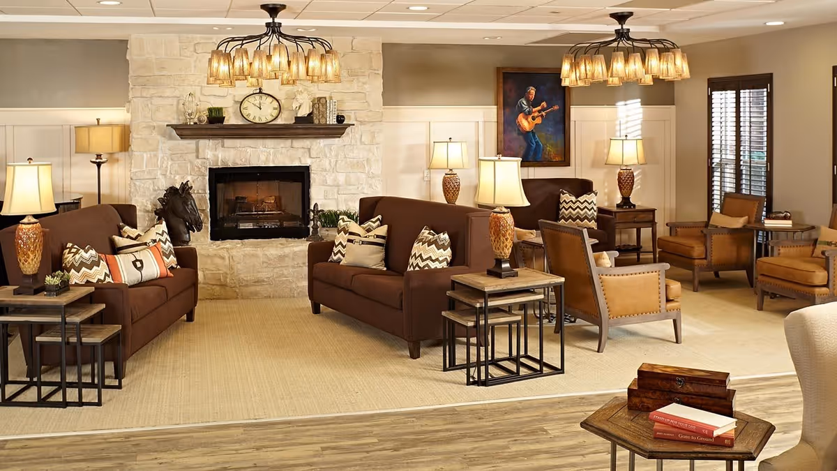 A cozy senior living facility lounge area with two brown sofas adorned with patterned cushions facing a stone fireplace. The room features warm lighting from multiple table lamps and two ceiling chandeliers. There are also several brown leather armchairs, side tables, a painting of a man playing guitar on the wall, and wooden flooring with a large beige rug.