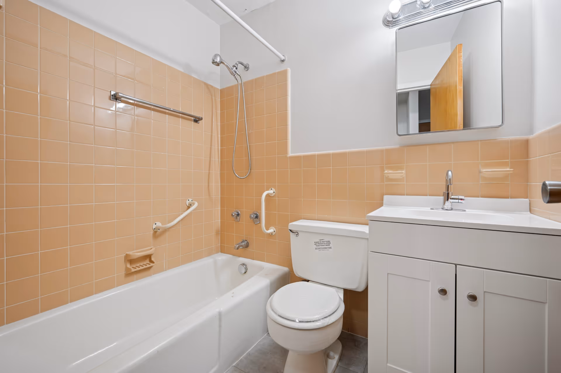 A bathroom with peach-colored tiled walls, a white bathtub with grab bars, a showerhead, a white toilet, and a white vanity with a sink and mirror above it.