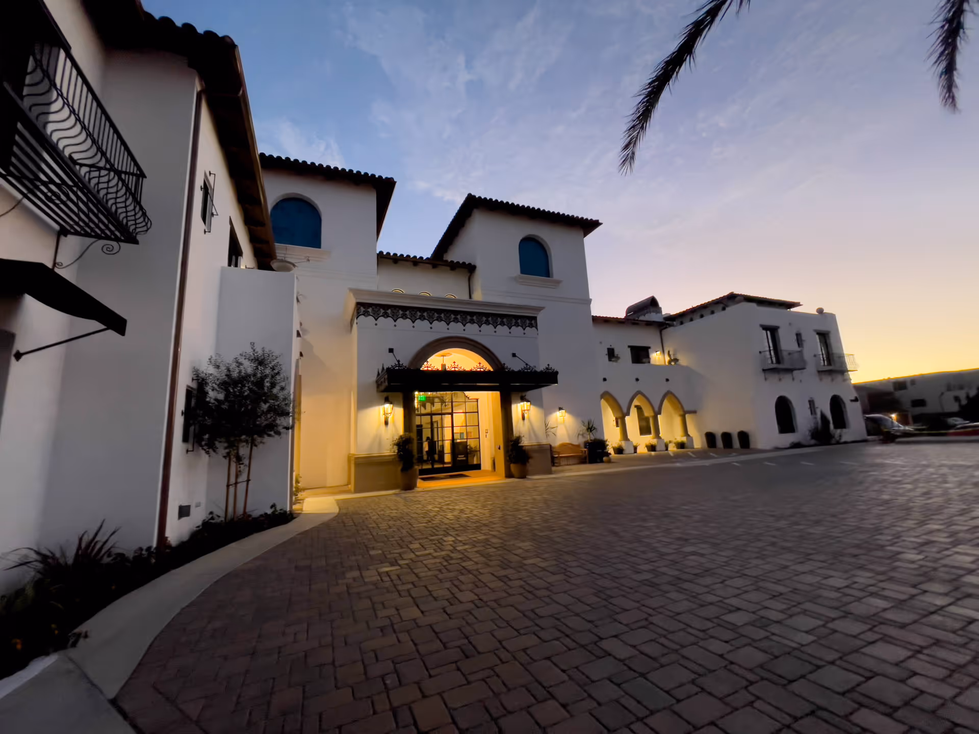 Mediterranean-style senior living building entrance at dusk with an illuminated doorway and brick driveway.