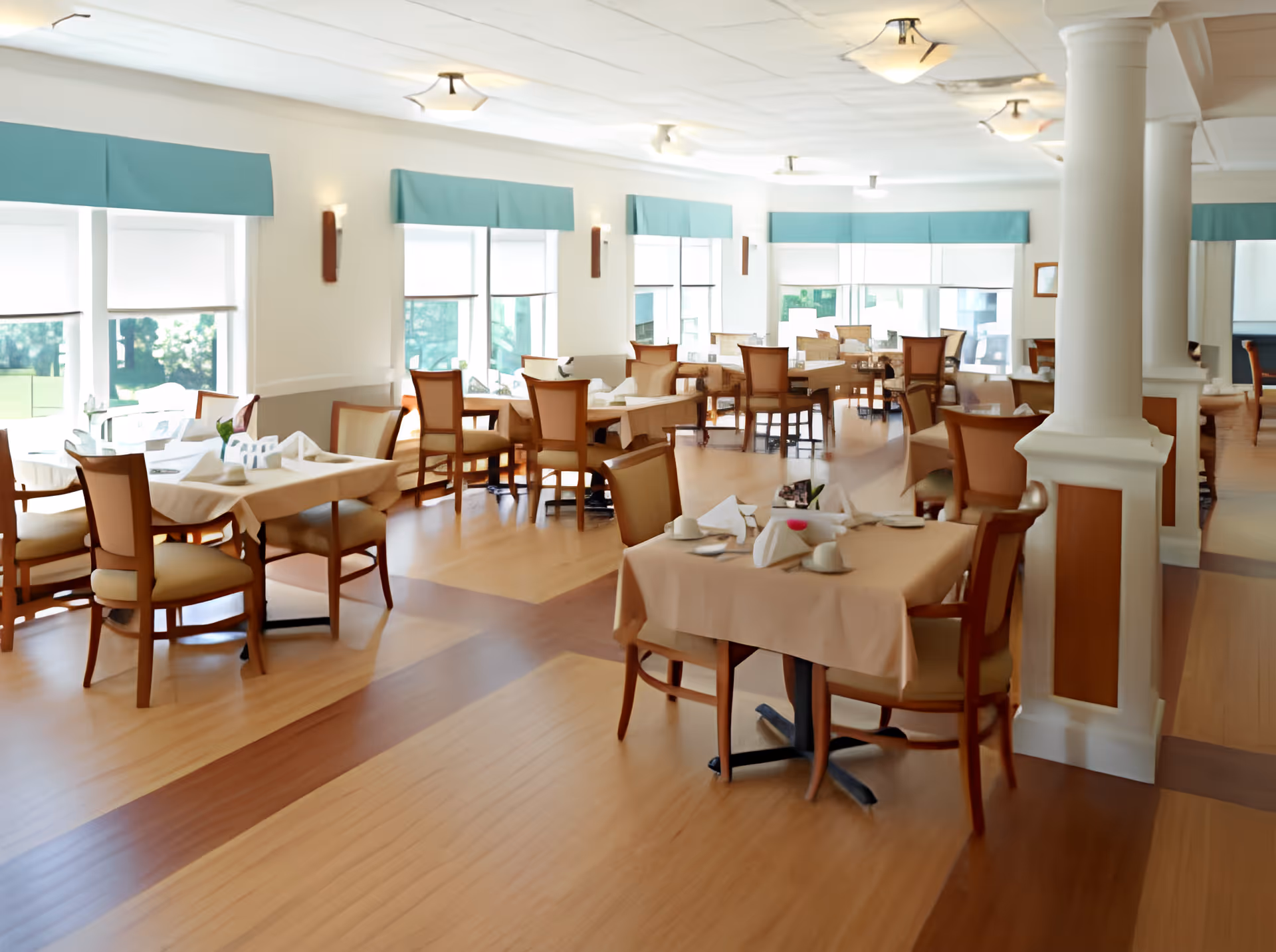 A bright and spacious dining room with multiple tables covered in beige tablecloths, each set with napkins and tableware. The room features large windows with teal valances, wooden chairs with cushioned seats, and a polished wooden floor. White pillars and soft ceiling lights add to the elegant and welcoming atmosphere.