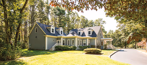 A large, light gray residential-style building with multiple dormer windows and a dark shingled roof, surrounded by green grass and trees under a clear sky.