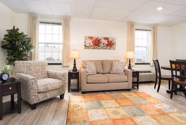 A bright living room with two large windows covered by beige curtains. The room features a beige loveseat with two patterned pillows, a matching armchair with script design, two dark wooden side tables each with a lamp, a colorful patterned area rug, a round dining table with four chairs, and a large green potted plant in the corner.