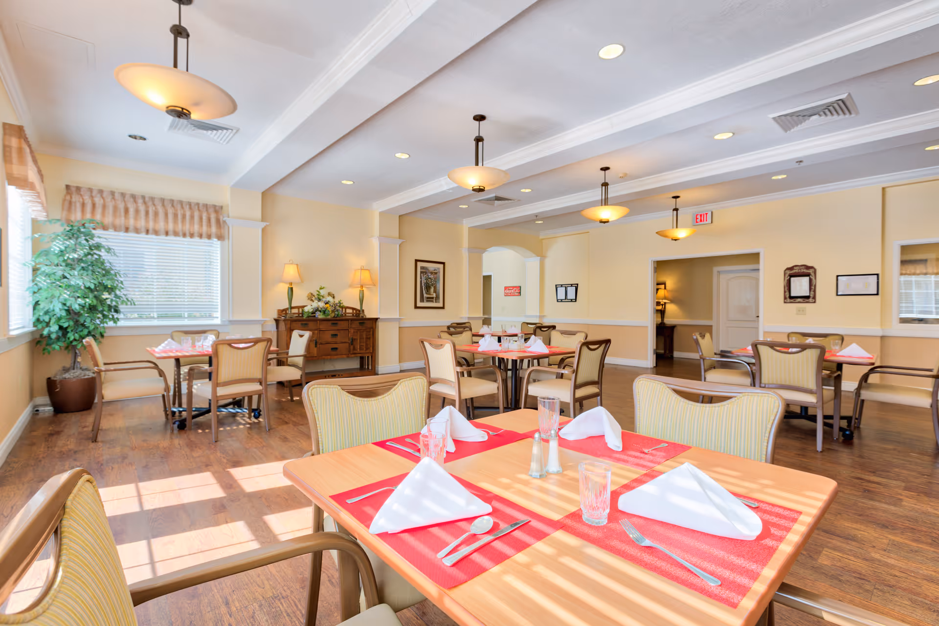 Bright dining room with several tables set with red placemats, white folded napkins, glasses, and silverware. The room has wooden floors, cream-colored walls, ceiling lights, and large windows with blinds allowing natural light. There is a potted plant near the window and a wooden sideboard with lamps and a floral arrangement against the wall.
