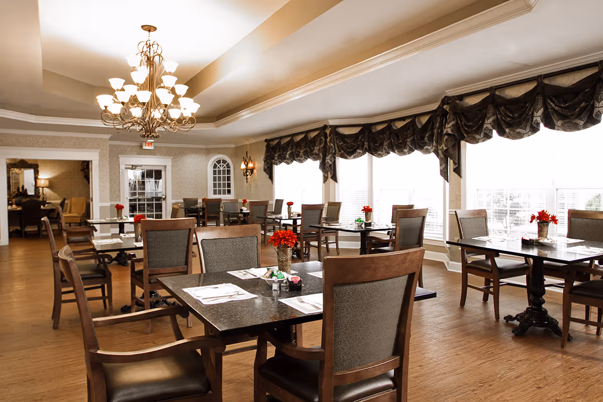 Bright, elegant dining room with multiple tables and chairs, chandeliers, large windows with draped valances, and red floral centerpieces.