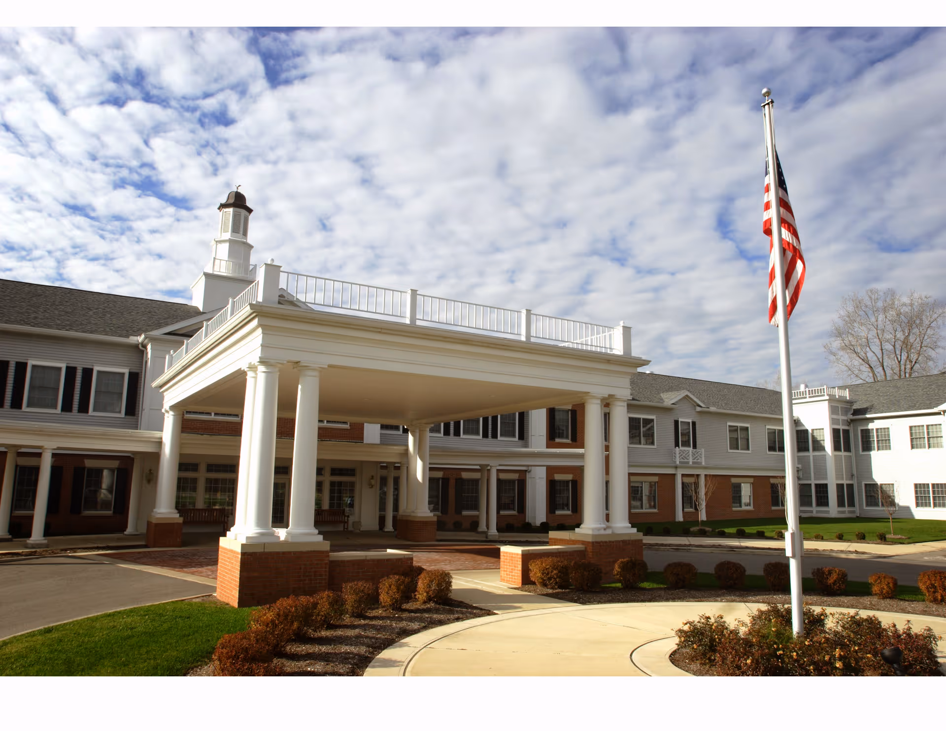 Exterior view of Kingston Residence of Sylvania, showing a large building with white columns supporting a covered entrance, an American flag on a flagpole, and partly cloudy sky.