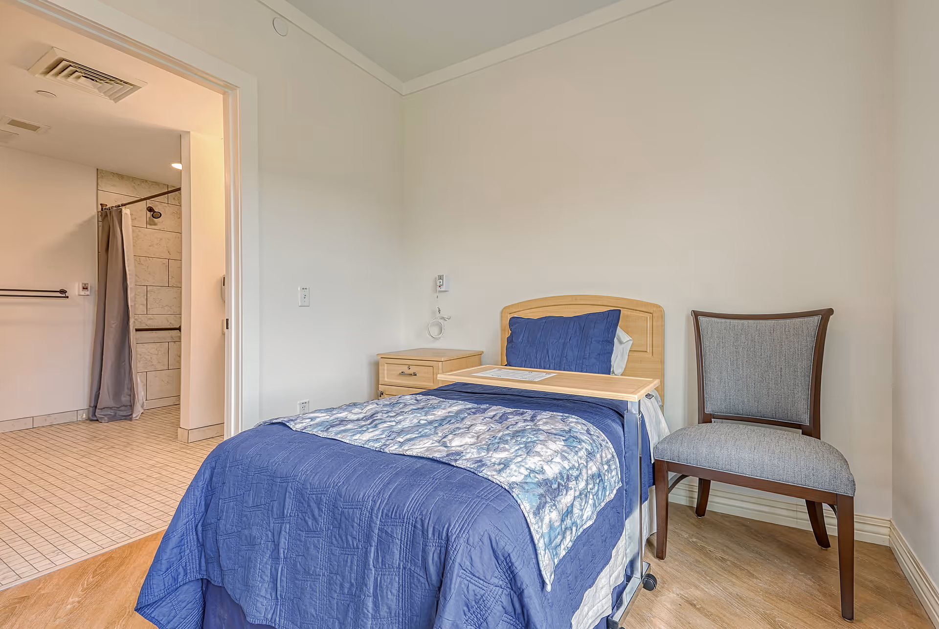 A small bedroom with a single bed covered in blue bedding and a patterned blanket. Next to the bed is a wooden nightstand and a gray upholstered chair. An overbed table is positioned over the bed. The room has light-colored walls and wood flooring. An open door reveals a bathroom with tiled floors and walls, a shower with a curtain, and grab bars.