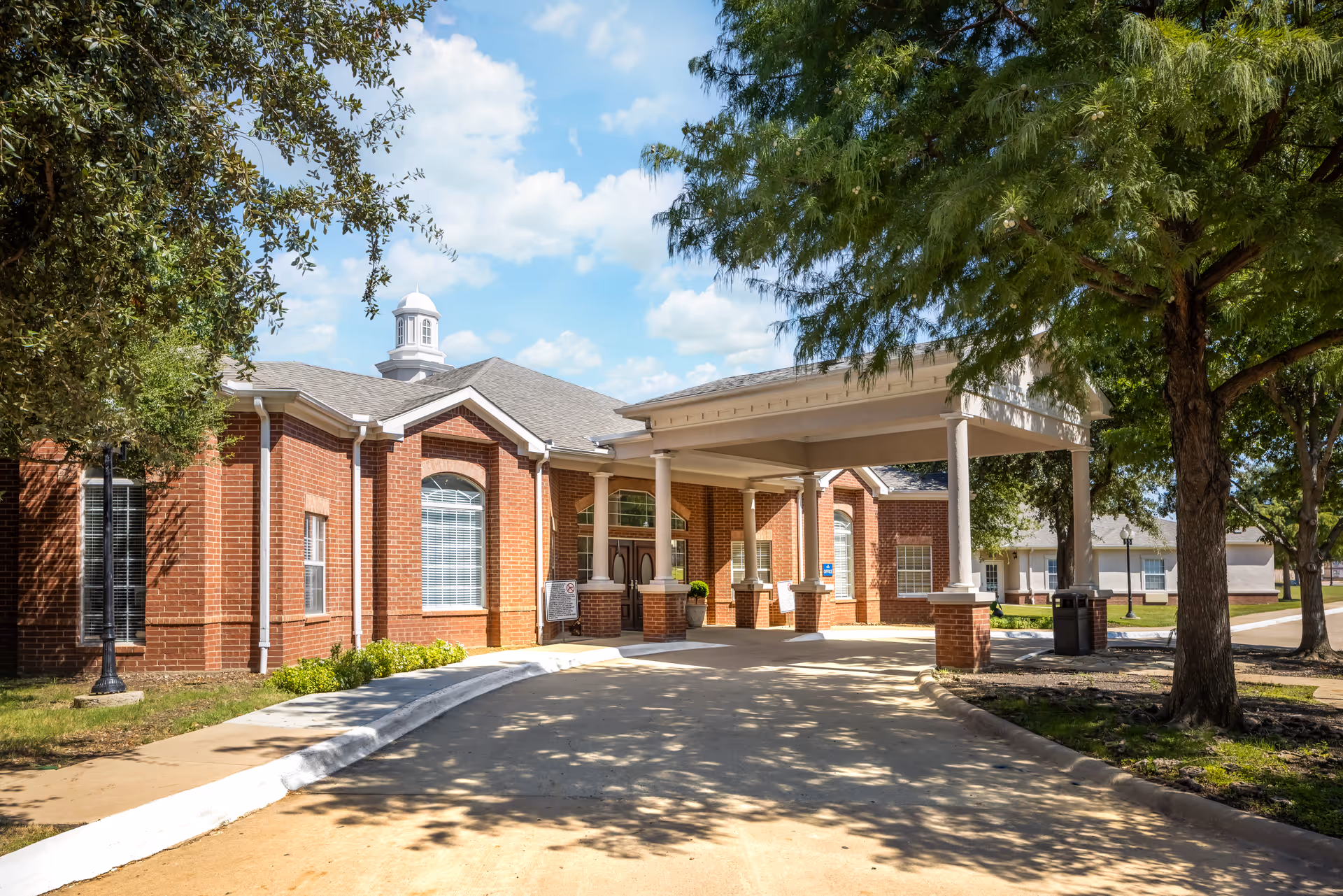 Exterior view of a brick building with a covered entrance supported by columns, surrounded by trees and a clear blue sky with some clouds.