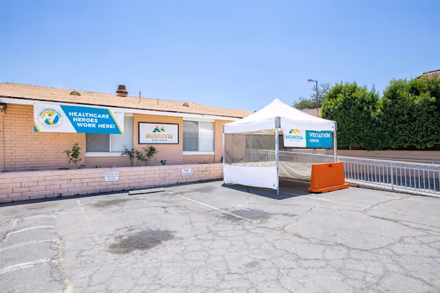 Outdoor parking area in front of a single-story brick building with signs for Monrovia Post Acute and a Healthcare Heroes Work Here banner. A white tent labeled Visitation Center is set up in the parking lot next to the building.