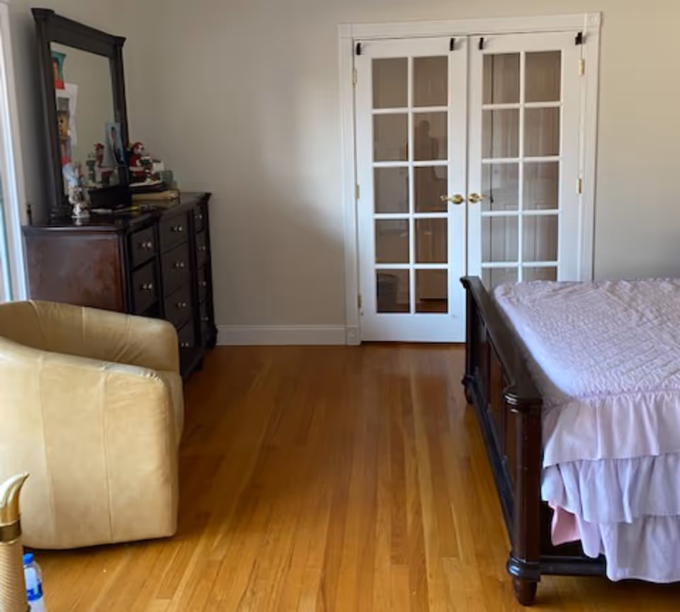 Bedroom with hardwood floors, a wooden bed with pink bedding, a dresser with mirror, a beige armchair, and white French doors.