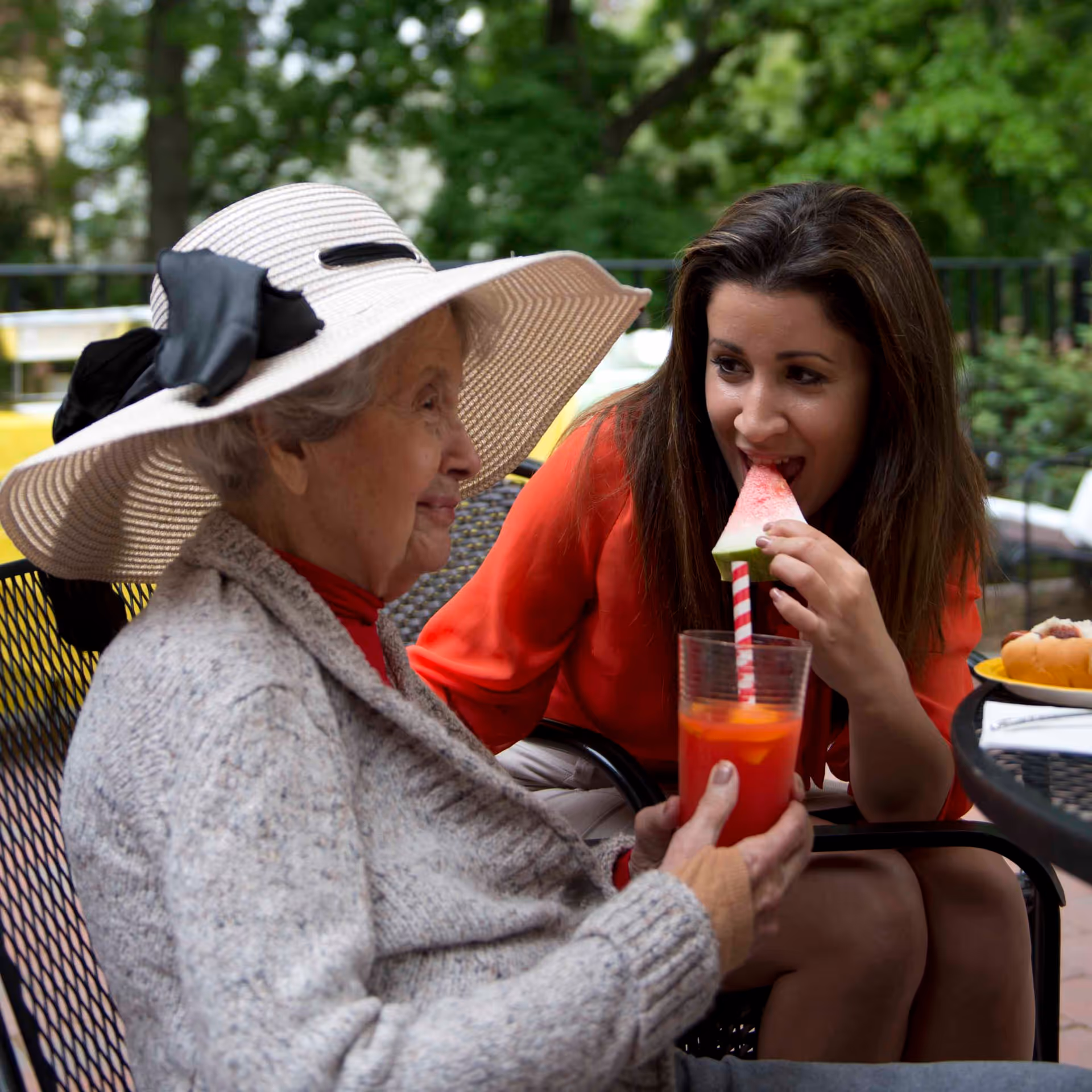 An elderly woman wearing a large white hat and gray sweater sits outdoors holding a glass of red drink with a striped straw. A younger woman in a red top is sitting next to her, eating a slice of watermelon. They are seated at a black metal table with a hot dog and napkins visible on it, surrounded by greenery.