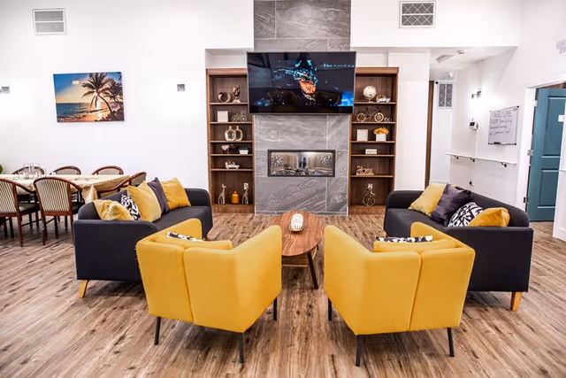 A cozy senior living common area with two dark gray sofas and two yellow armchairs arranged around a wooden coffee table. A modern fireplace is set in a gray tiled wall with built-in wooden shelves on either side, decorated with various ornaments. A flat-screen TV is mounted above the fireplace. To the left, there is a dining table with chairs, and a wall-mounted picture of a palm tree at sunset. The floor is wood, and the walls are white.