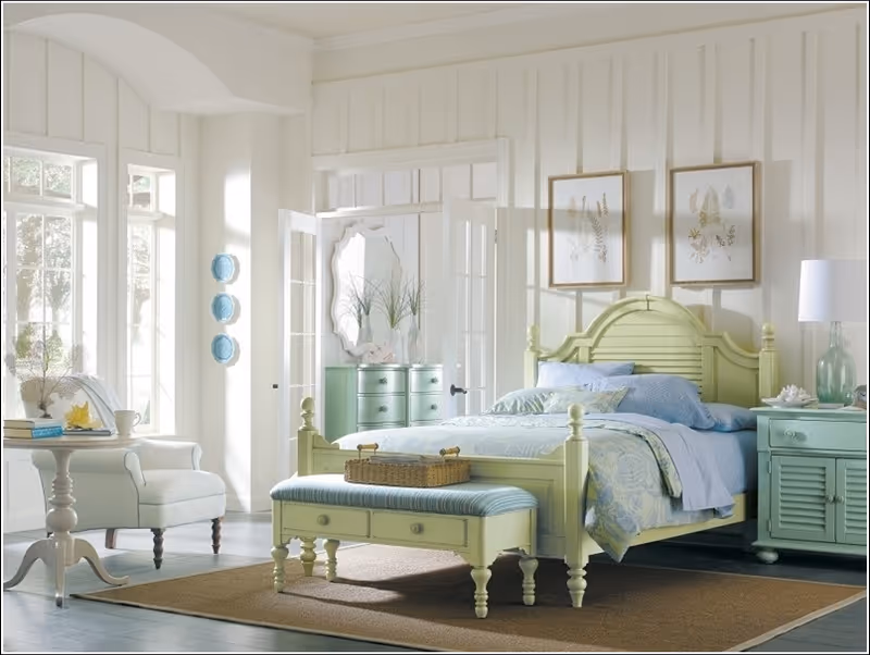 A bright and airy bedroom with a light green wooden bed frame and matching bench at the foot of the bed. The bed is made with blue and green bedding. There is a nightstand with a lamp and decorative items on the right side of the bed. On the left side, there is a white armchair next to a small round table with books and a cup. The room has large windows letting in natural light, white paneled walls, and two framed botanical prints above the bed. In the background, a dresser with a mirror and plants is visible through an open doorway.