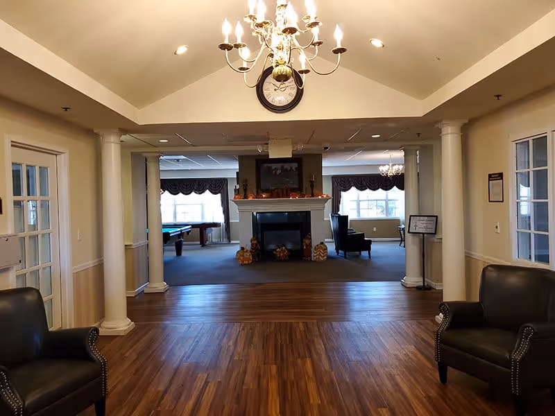 Interior view of a senior living facility common area featuring a wooden floor leading to a carpeted lounge with a fireplace decorated for autumn. Two black leather armchairs are positioned on either side of the wooden floor area. White columns frame the entrance to the lounge, which has large windows with valances, a chandelier hanging from the ceiling, and a clock mounted above the fireplace.
