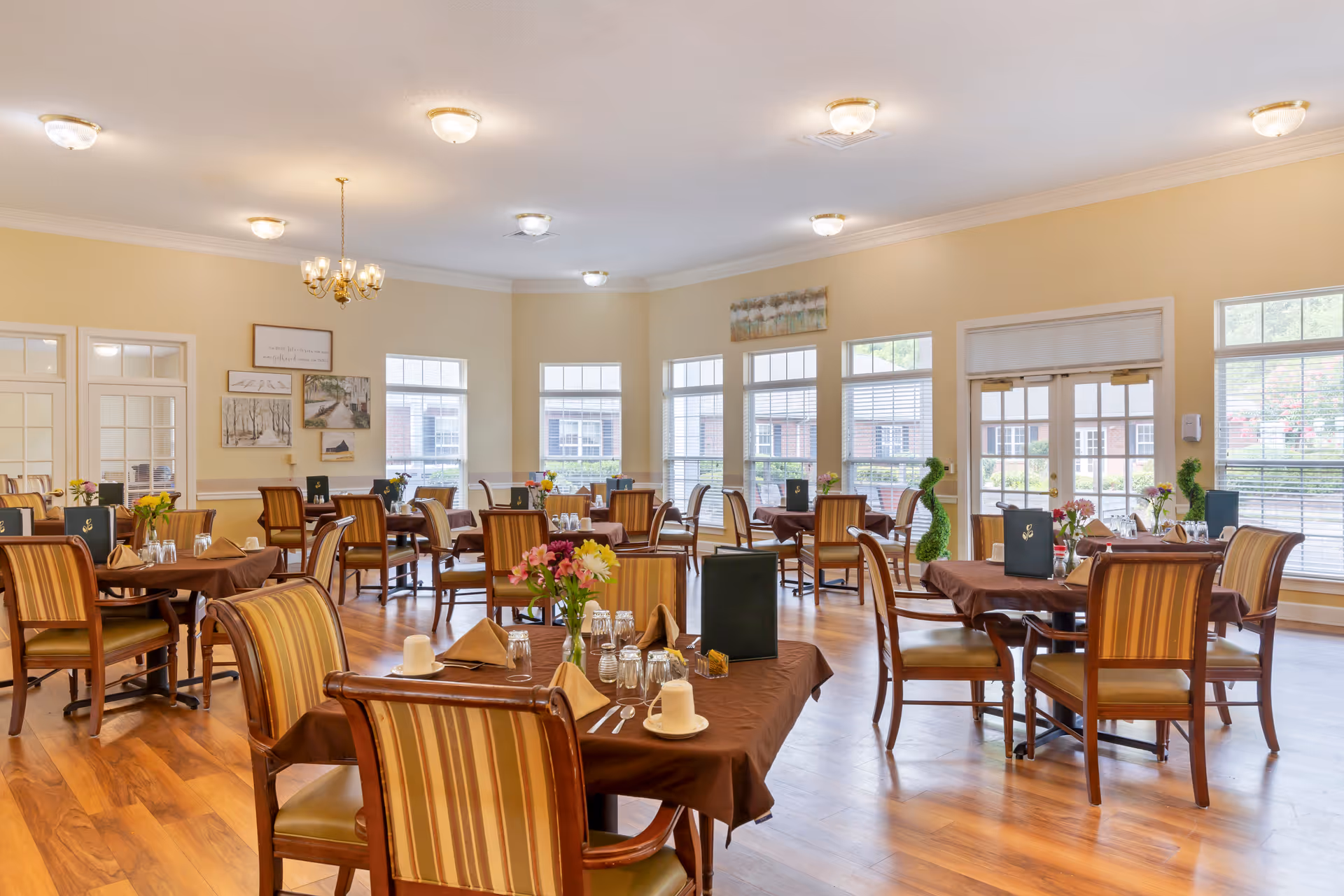 A bright and spacious dining room with multiple tables covered in brown tablecloths, each set with napkins, glasses, cups, and small flower arrangements. The room has large windows allowing natural light to fill the space, wooden floors, and beige walls decorated with framed artwork. Chairs with striped upholstery surround the tables.