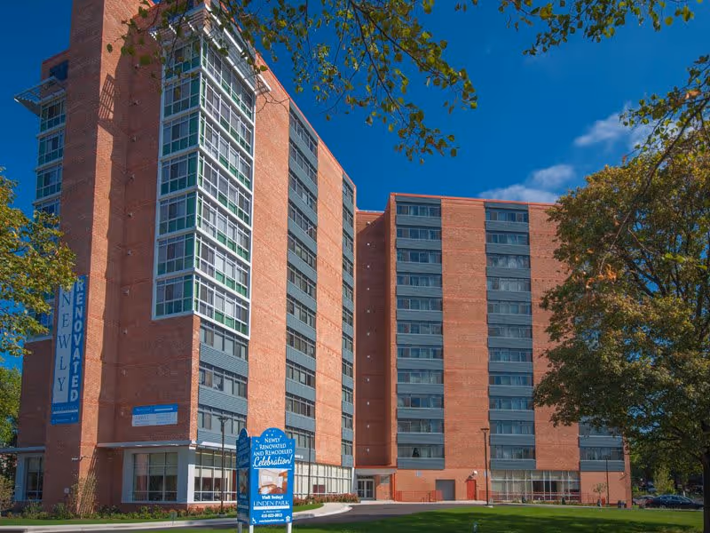 Exterior view of a tall brick apartment building under a clear blue sky with some trees around. The building has multiple floors with large windows and a sign in front that reads 'Newly Renovated and Remodeled Celebration' for Linden Park Apartments.