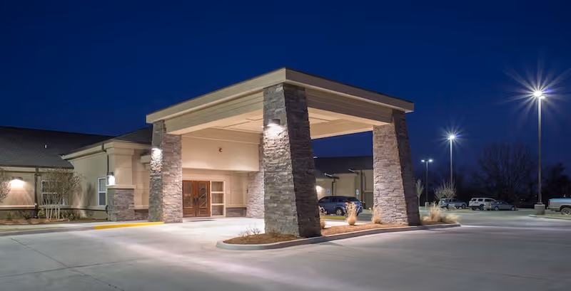 Exterior nighttime view of the entrance to Cherokee County Nursing Center, featuring a covered drop-off area supported by stone pillars, illuminated by outdoor lights with a parking lot and several vehicles in the background.