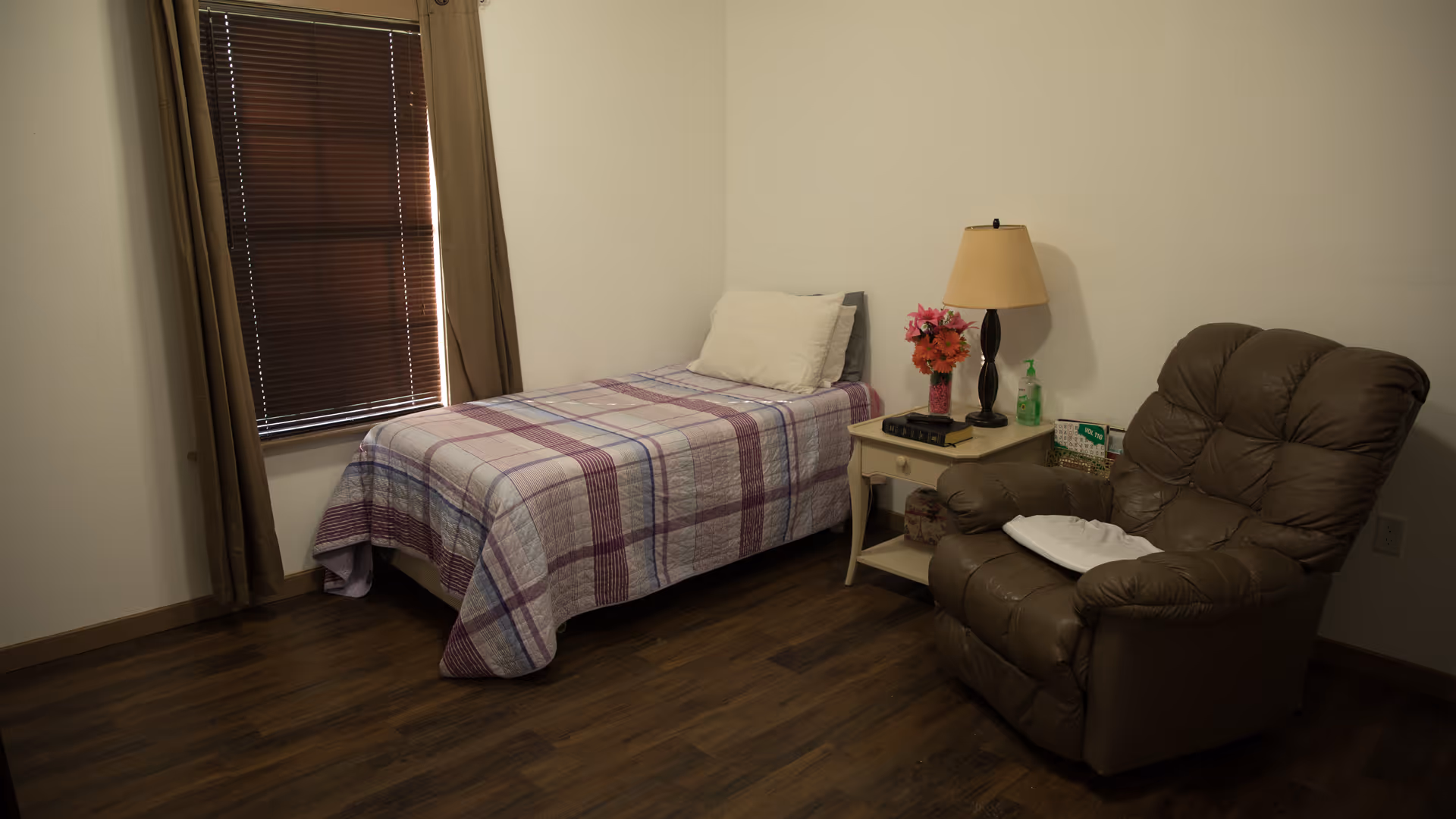 A small bedroom with a single bed covered in a plaid quilt, a pillow, a nightstand with a lamp, flowers, and hand sanitizer, and a brown recliner chair. The room has wooden flooring and a window with closed brown blinds and curtains.