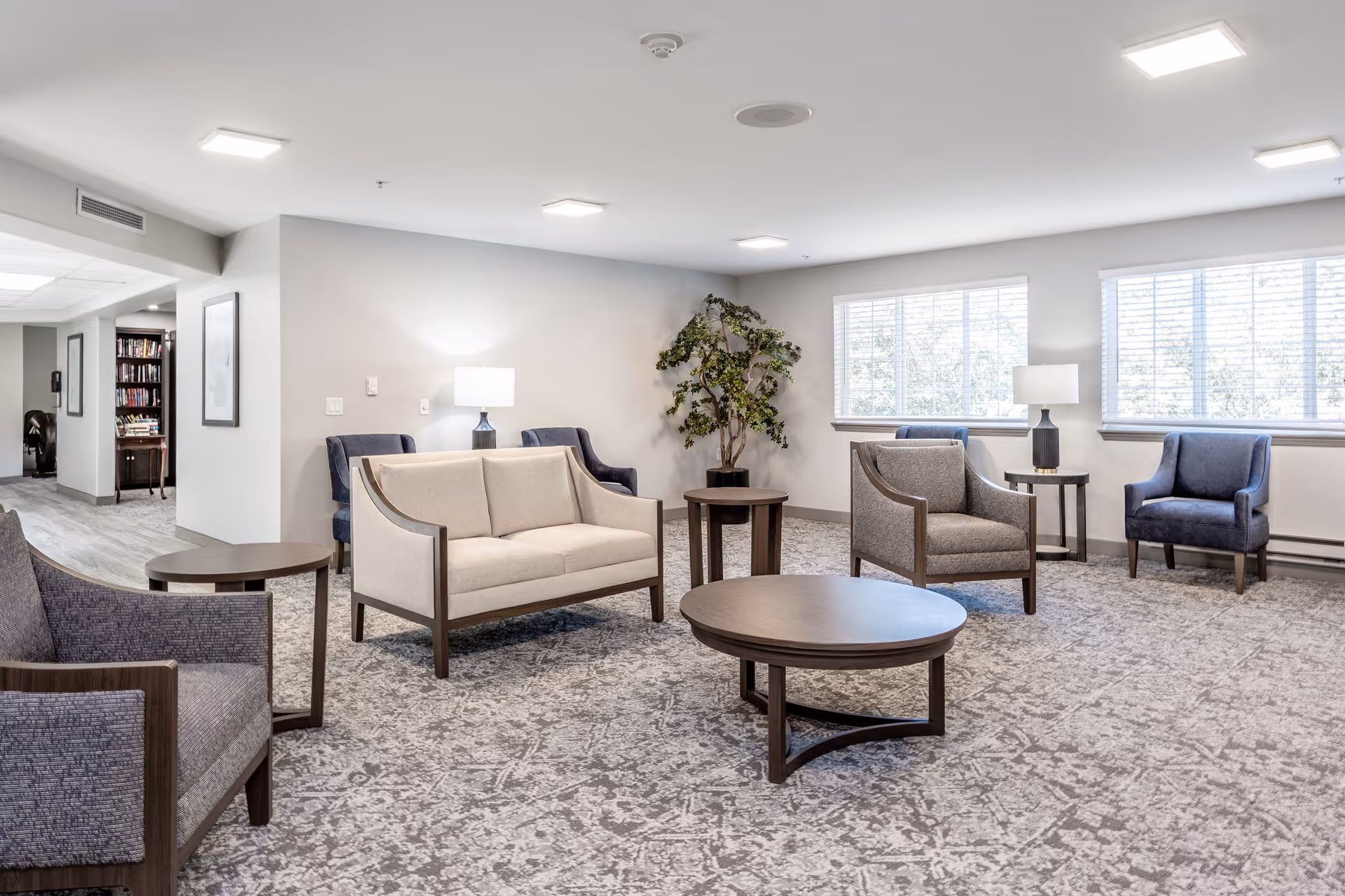 Bright, spacious senior living lounge with sofas, armchairs, coffee tables, lamps, and large windows.
