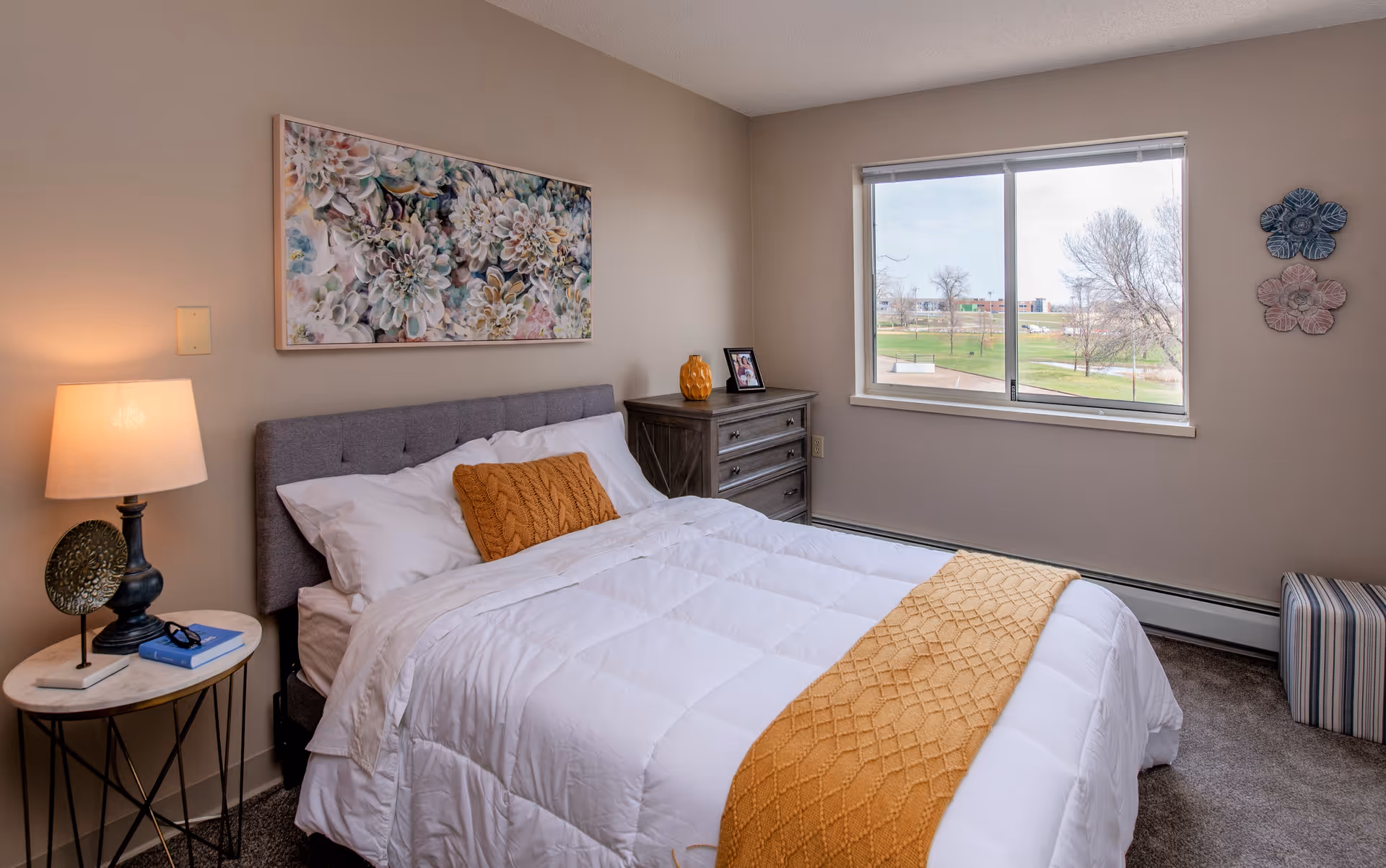 A cozy bedroom with a neatly made bed featuring white bedding and a mustard yellow throw blanket and pillow. There is a bedside table with a lamp, a small decorative item, and a book. A dresser with a framed photo and a decorative vase is against the wall. A large window shows an outdoor view with trees and a grassy area. The walls are beige with floral artwork above the bed and three decorative flower wall hangings near the window.