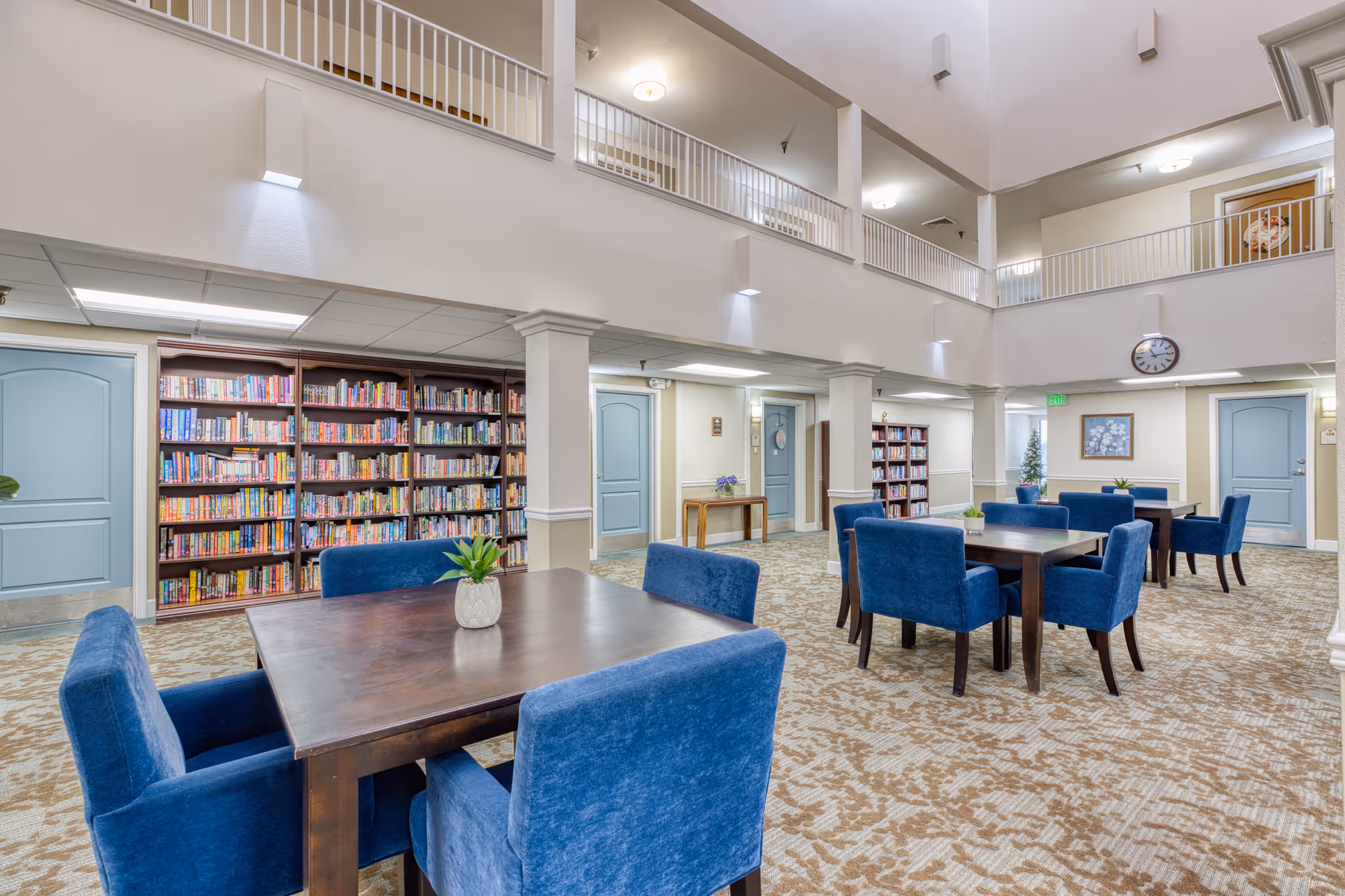 A spacious, well-lit common area with blue upholstered chairs around dark wooden tables, beige patterned carpet, and bookshelves filled with books along the walls. The room has a high ceiling with a second-floor balcony overlooking the area, light blue doors, a wall clock, and framed artwork.