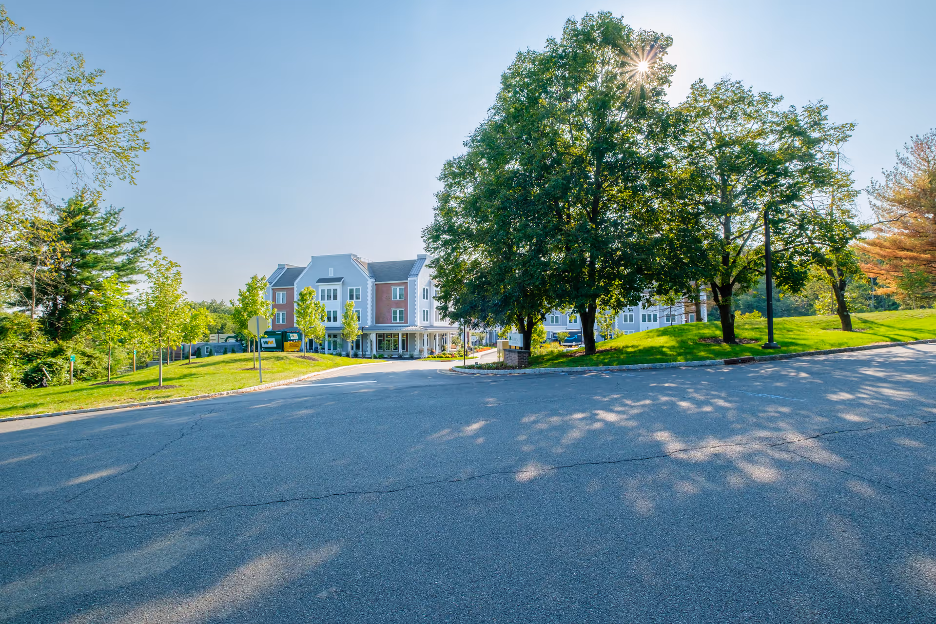 Exterior view of The Residence at Basking Ridge senior living facility on a sunny day, showing a paved driveway, green grass, several trees, and a multi-story building with a porch and multiple windows.