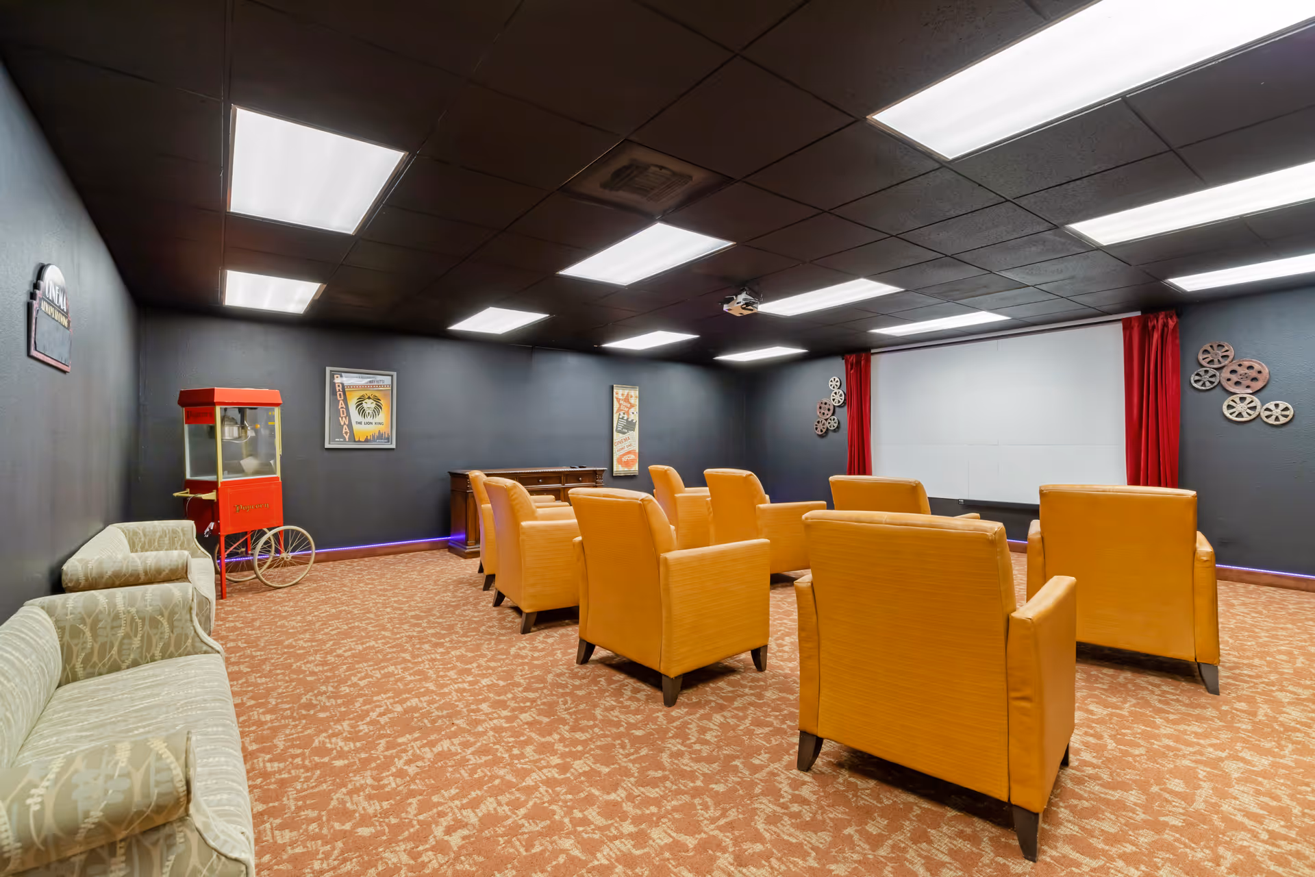 A small movie theater room with several orange armchairs arranged in rows facing a large white screen with red curtains on the sides. The room has dark walls decorated with vintage movie posters and film reels. There is a popcorn machine and a beige patterned couch along the left wall. The ceiling has multiple rectangular fluorescent lights.