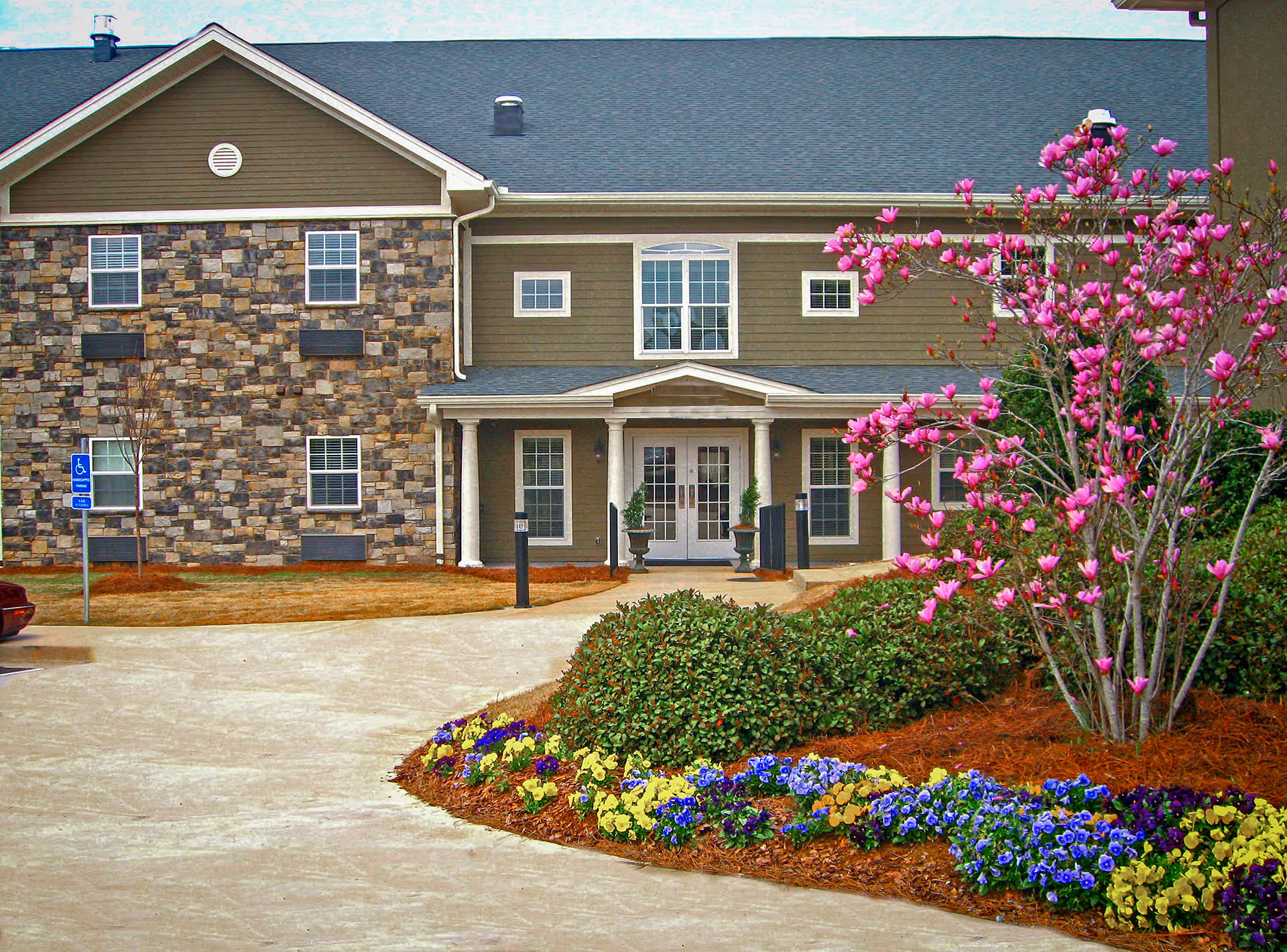 Exterior view of Benton Village of Stockbridge showing a two-story building with stone and siding facade, a covered entrance with white columns, a driveway, and landscaped flower beds with colorful flowers and a blooming pink tree.