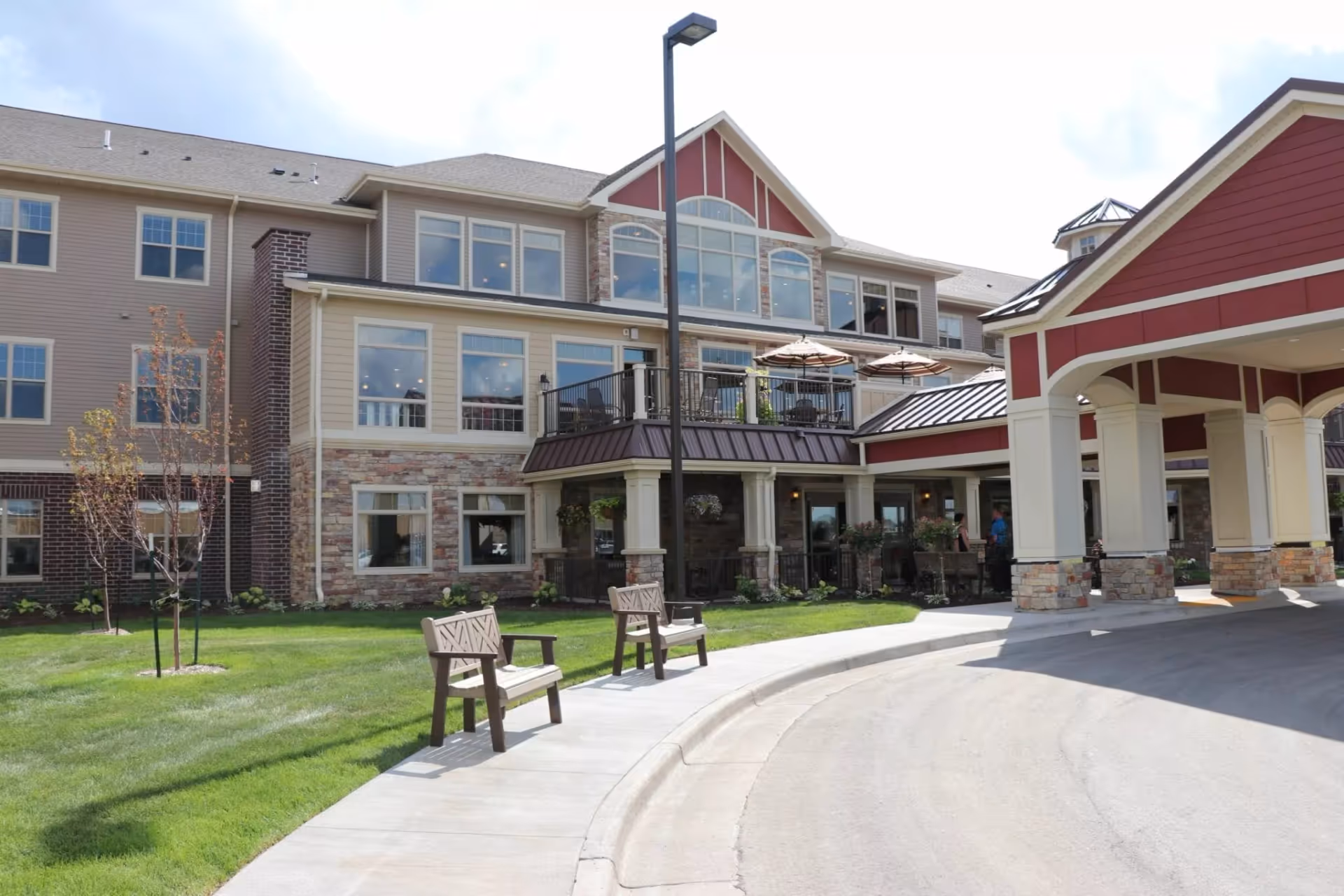 Exterior view of New Perspective Senior Living facility in West Fargo showing a multi-story building with large windows, a covered entrance with stone pillars, outdoor benches along a curved sidewalk, and a well-maintained lawn with small trees.
