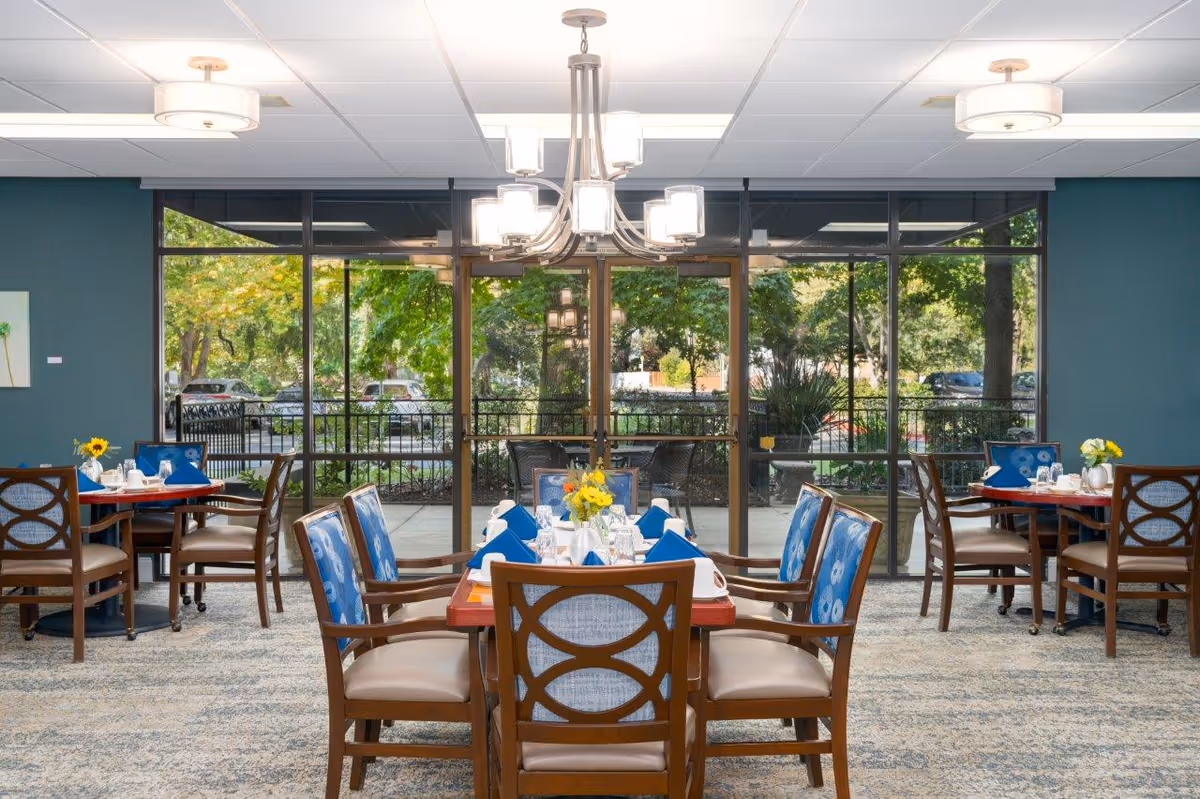 A bright dining room with several wooden tables and chairs arranged neatly. Each table is set with white tableware, blue napkins, and small flower centerpieces. Large floor-to-ceiling windows provide a view of green trees and parked cars outside. A modern chandelier hangs from the ceiling, illuminating the room.