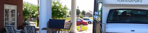 Outdoor view of a covered porch area with chairs and a grill, adjacent to a brick building. A white vehicle labeled 'Plantation Pointe' is parked nearby, with a red car and greenery visible in the background.