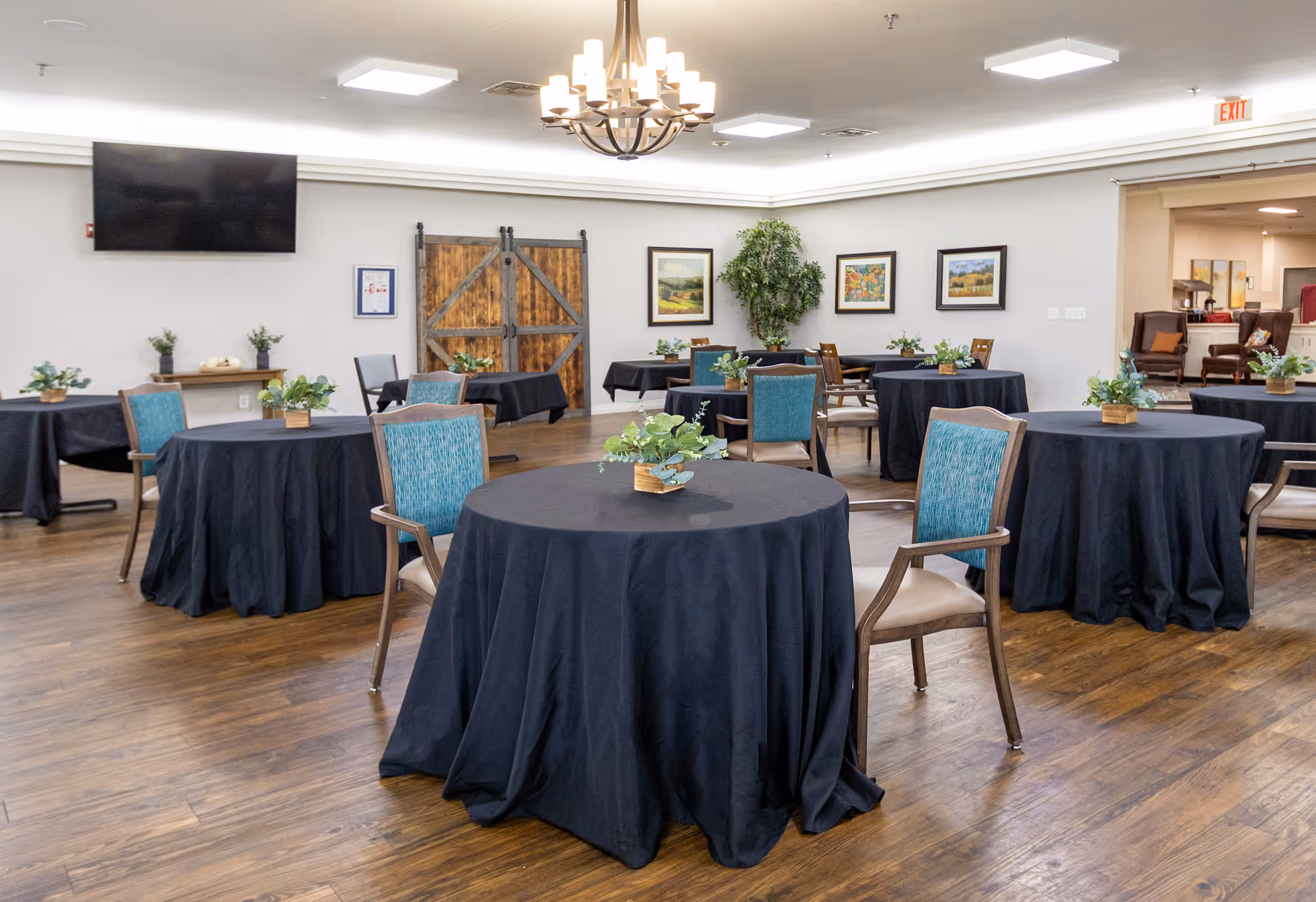 A spacious dining room with round tables covered in black tablecloths, each adorned with small green potted plants. The room has wooden flooring, teal cushioned chairs with wooden frames, a chandelier hanging from the ceiling, framed artwork on the walls, and a large flat-screen TV mounted on one wall. There is a wooden barn-style door and an exit sign visible in the background.