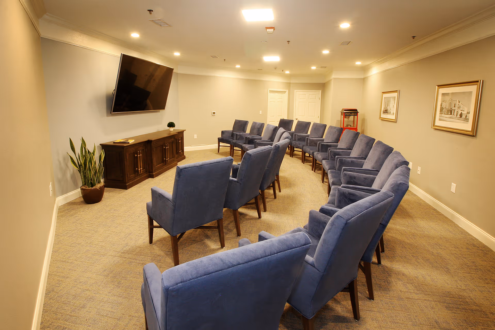 A senior living facility common room with two rows of blue upholstered chairs arranged facing a large wall-mounted flat screen TV. The room has beige walls, carpeted floor, a wooden cabinet under the TV, a potted plant, framed pictures on the walls, and a popcorn machine in the back corner.