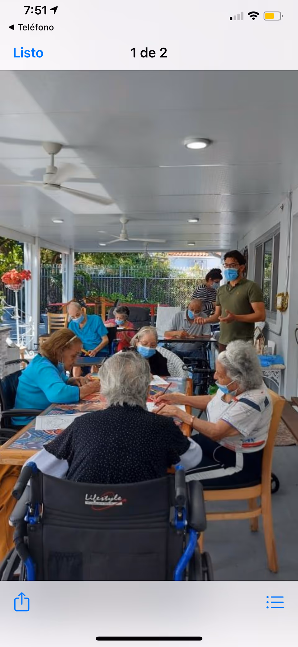 A group of elderly people wearing face masks sitting around tables on a covered outdoor patio, engaging in activities with a caregiver standing nearby also wearing a mask. The area is well-lit with ceiling fans and surrounded by greenery.