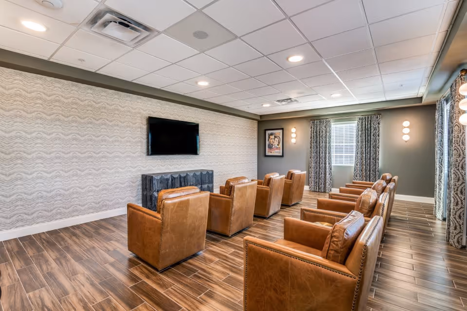 A senior living facility common room with multiple brown leather armchairs arranged in rows facing a wall-mounted flat screen TV. The room has wood-look tile flooring, patterned wallpaper on one wall, and windows with patterned curtains. Soft wall lights and ceiling lights illuminate the space.