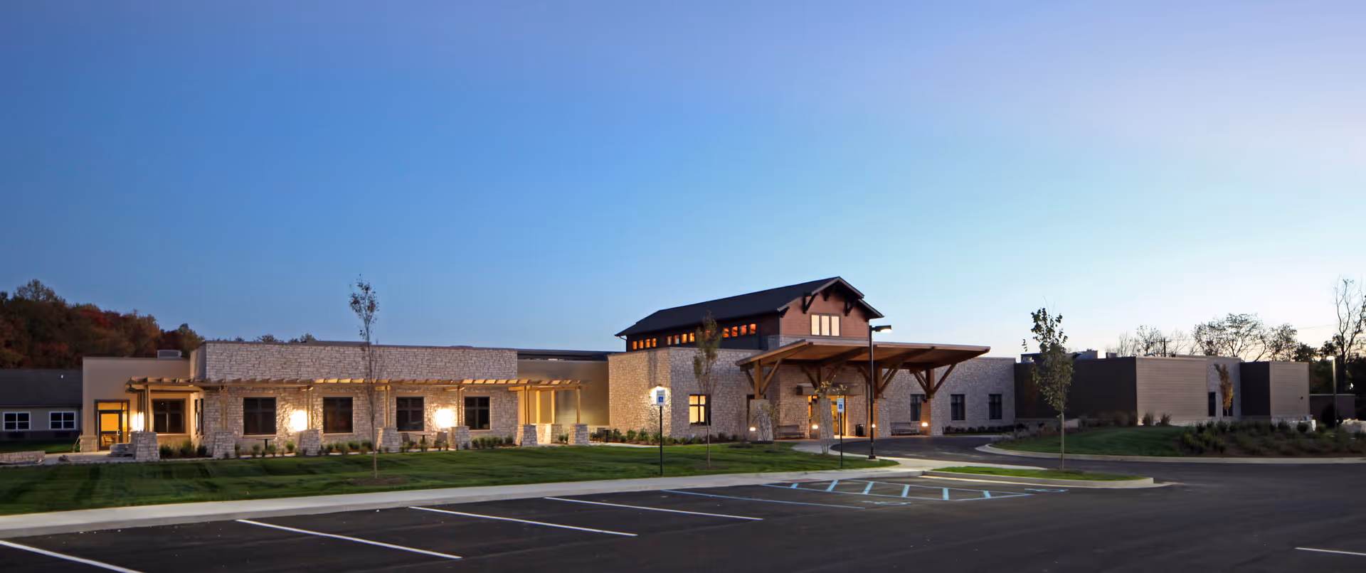 Exterior view of Wellbrooke of Crawfordsville facility during dusk, showing a modern single-story building with stone and beige walls, large windows, a covered entrance with wooden beams, and an empty parking lot in the foreground.
