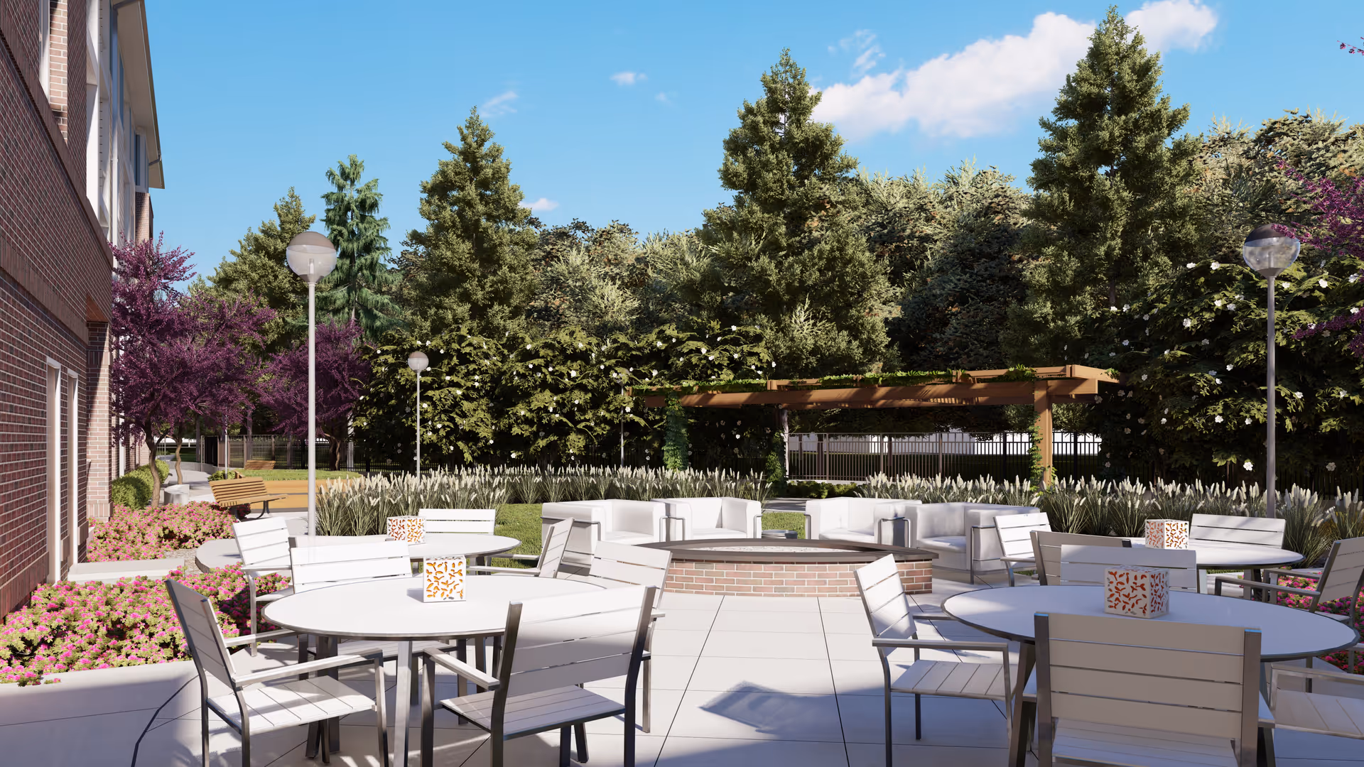 Outdoor patio area with round white tables and chairs arranged around a central brick fire pit. The patio is surrounded by landscaped greenery including purple flowering trees, bushes, and tall grasses. There is a wooden pergola in the background and several lamp posts providing lighting. The sky is clear and blue.
