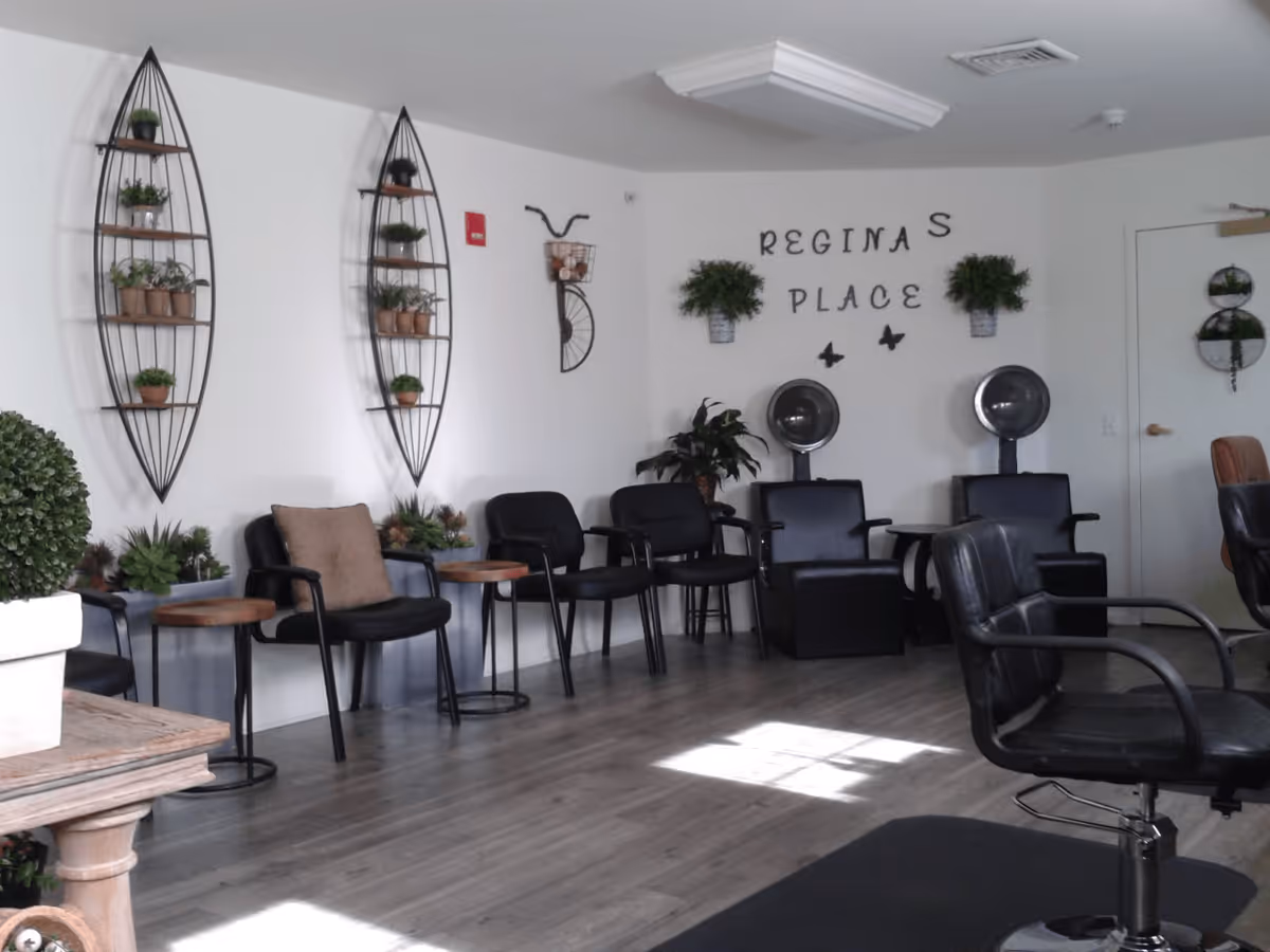Interior of a salon area with black chairs arranged along the walls and in the center, two hair dryer chairs, wall decorations including plants and a bicycle wall art, and the words 'REGINA'S PLACE' on the wall.