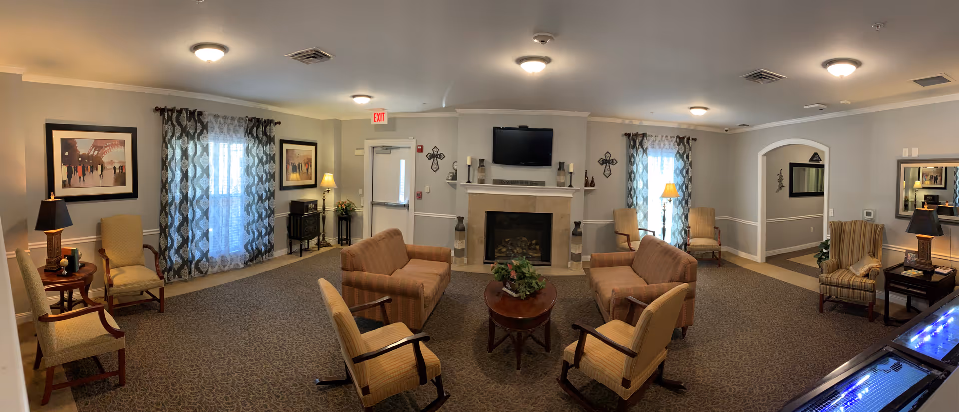 Warm senior living lounge with couches, chairs, fireplace, TV, and patterned curtains.