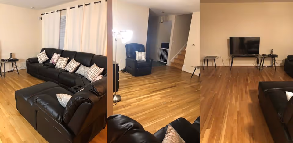 A spacious living room with light-colored walls and hardwood floors. The room features a large black leather sectional sofa with multiple patterned throw pillows, a matching black leather recliner chair with a pillow, a floor lamp, and a flat-screen TV on a stand against the wall. There are two small side tables near the sofa and TV. A staircase is visible in the background.