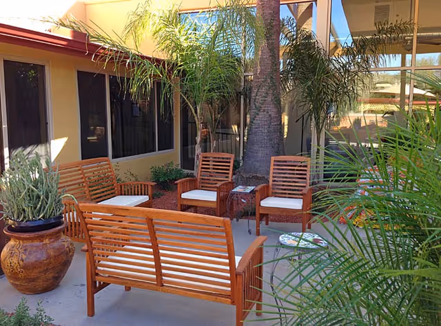 Outdoor patio area with wooden bench and chairs with white cushions arranged around a small table. The space is surrounded by potted plants and palm trees, adjacent to a building with large windows.