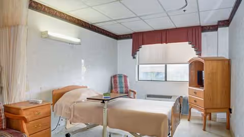 A small bedroom with a single bed, dresser, chair, and window in a senior facility.