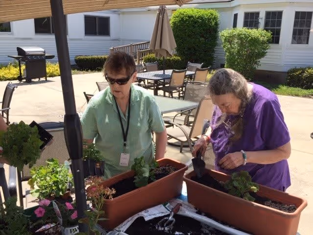 Two elderly women gardening outdoors at a senior living facility, planting green plants in rectangular planters on a table under a patio umbrella. There are patio chairs and tables in the background along with a barbecue grill and white building walls with windows.